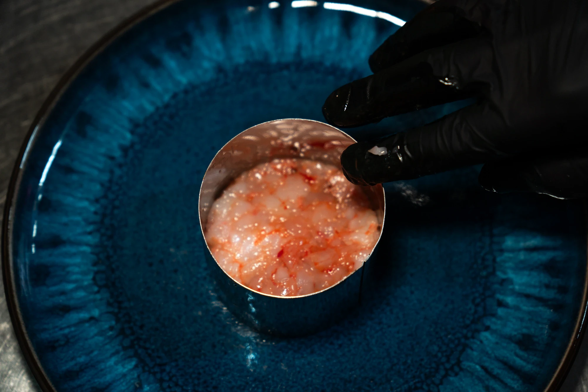 Mano con guanto nero che prepara una tartare di mare in uno stampo circolare su piatto blu