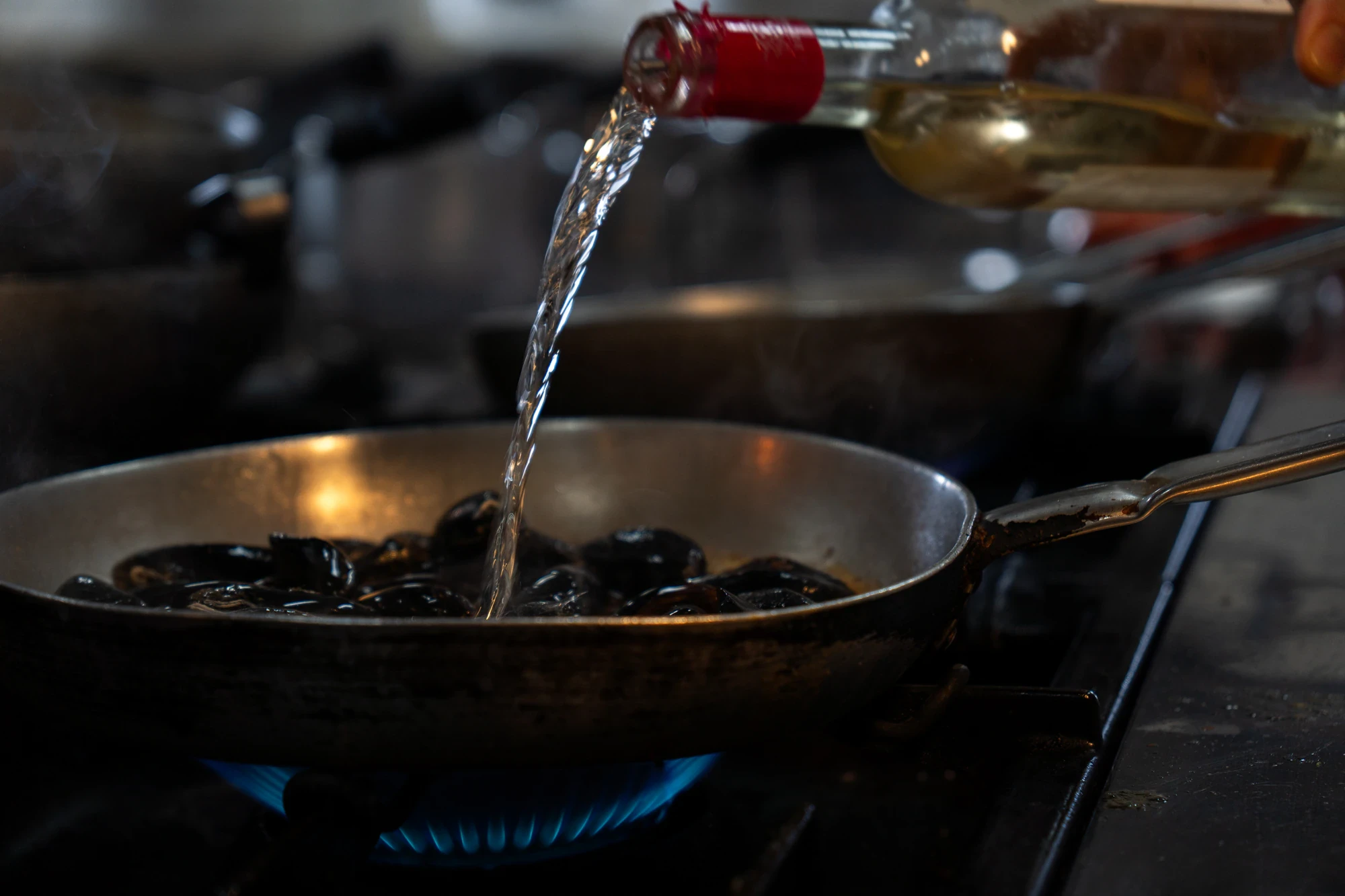 Versamento di vino bianco in una padella di cozze sul fuoco a gas blu